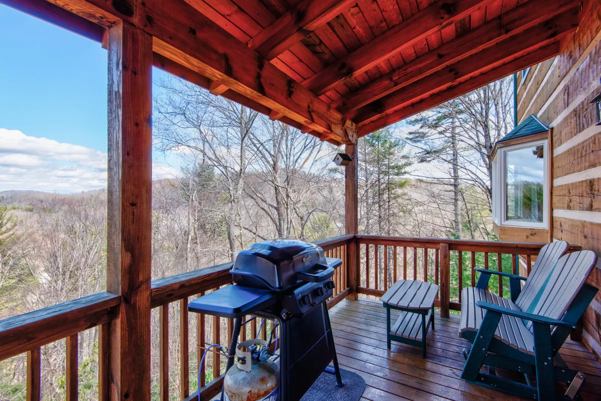 A wooden deck with a grill and Adirondack chairs sits above a canopy of trees, inviting outdoor dining and leisure. The view through the railing reveals vibrant foliage, suggesting an ideal spot for post-adventure gatherings or sunset watching.