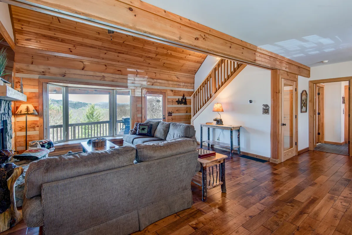 A spacious, open interior featuring a comfortable seating area with plush sofas, a prominent stone fireplace, and large windows that invite natural light and frame scenic views. The wooden staircase and cozy furniture make the space ideal for relaxing or entertaining.