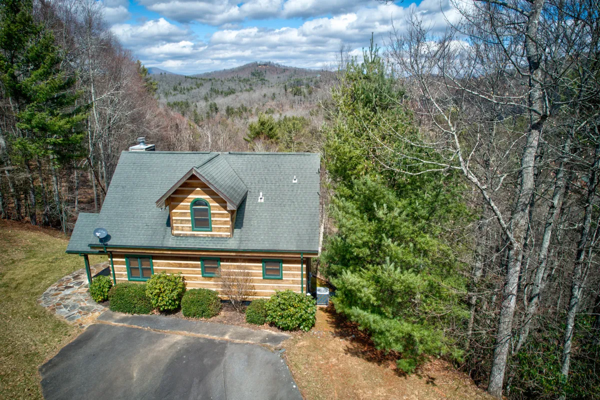 A classic log cabin sits nestled among tall trees, with a dramatic mountain view stretching beyond the treetops. The tranquil setting hints at early morning sunrises and the hush of evening, ideal for launching outdoor adventures or relaxing after a day’s exploration.