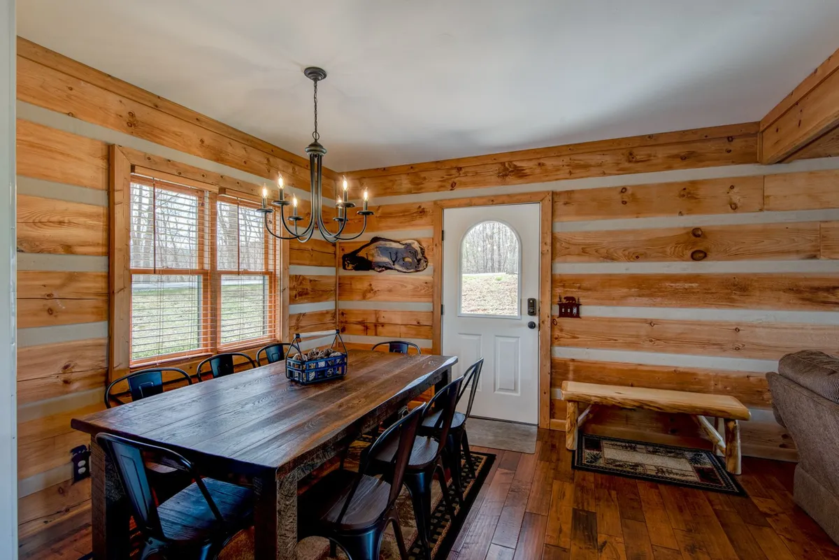 A large wooden dining table lit by a chandelier, flanked by windows that fill the room with natural light. The inviting setup suggests a welcoming atmosphere for family meals, celebrations, or game nights, with a door leading to other communal areas.