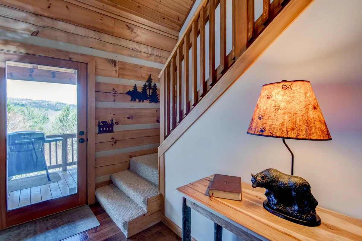 A lofted nook with a visible staircase, wooden railing, soft lamp light, bear figurine, and a book on the table. The space feels intimate and perfect for reading, reflection, or quiet work while overlooking the main living area.