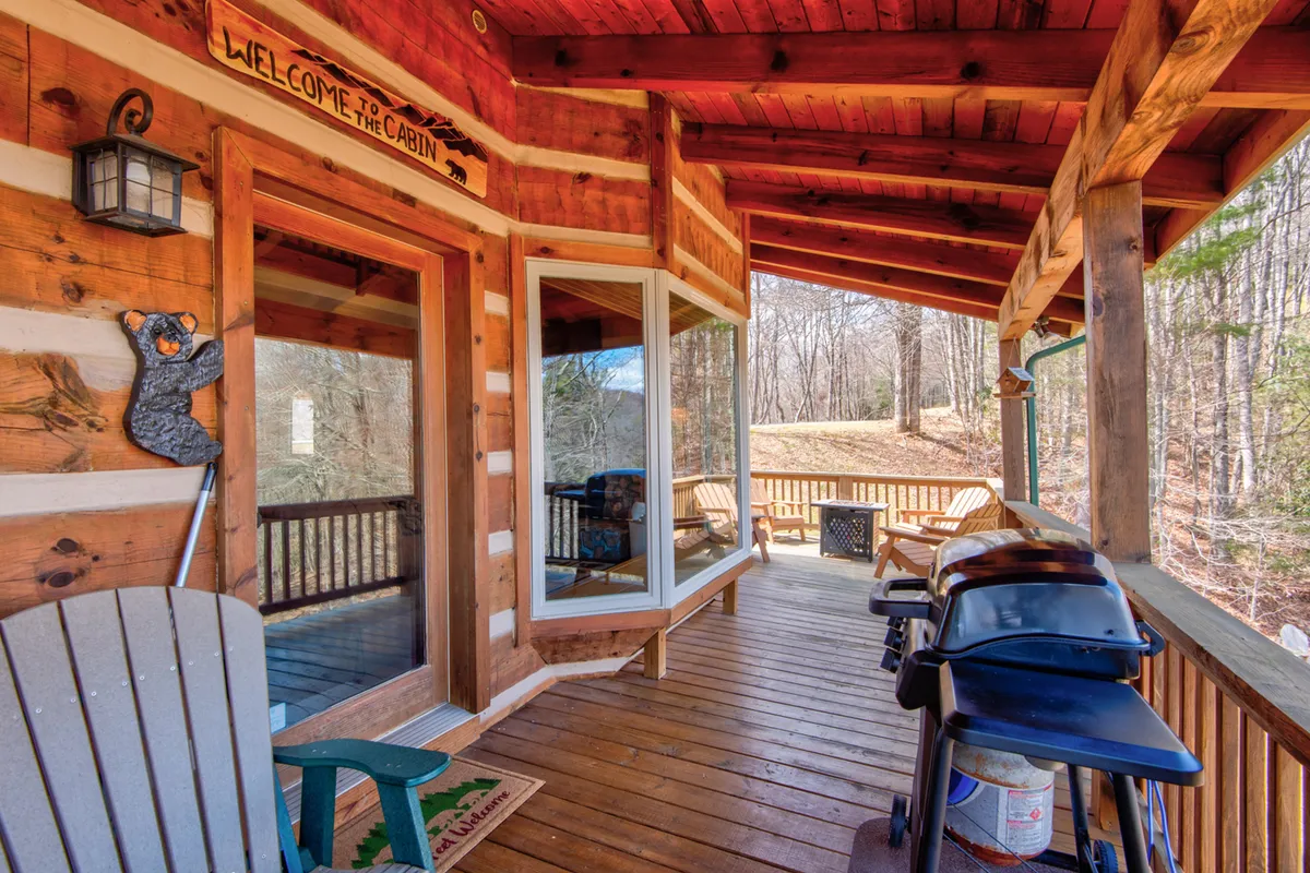 This exterior scene highlights the Cozy Bear Cabin's rustic log structure, welcoming porch with rocking chairs, grill, and a friendly 'welcome' sign. The warm lighting and natural surroundings evoke a sense of relaxation and invitation, ideal for couples or small groups seeking tranquility. Sunset-hued skies further enhance the sense of serenity and mountain charm.