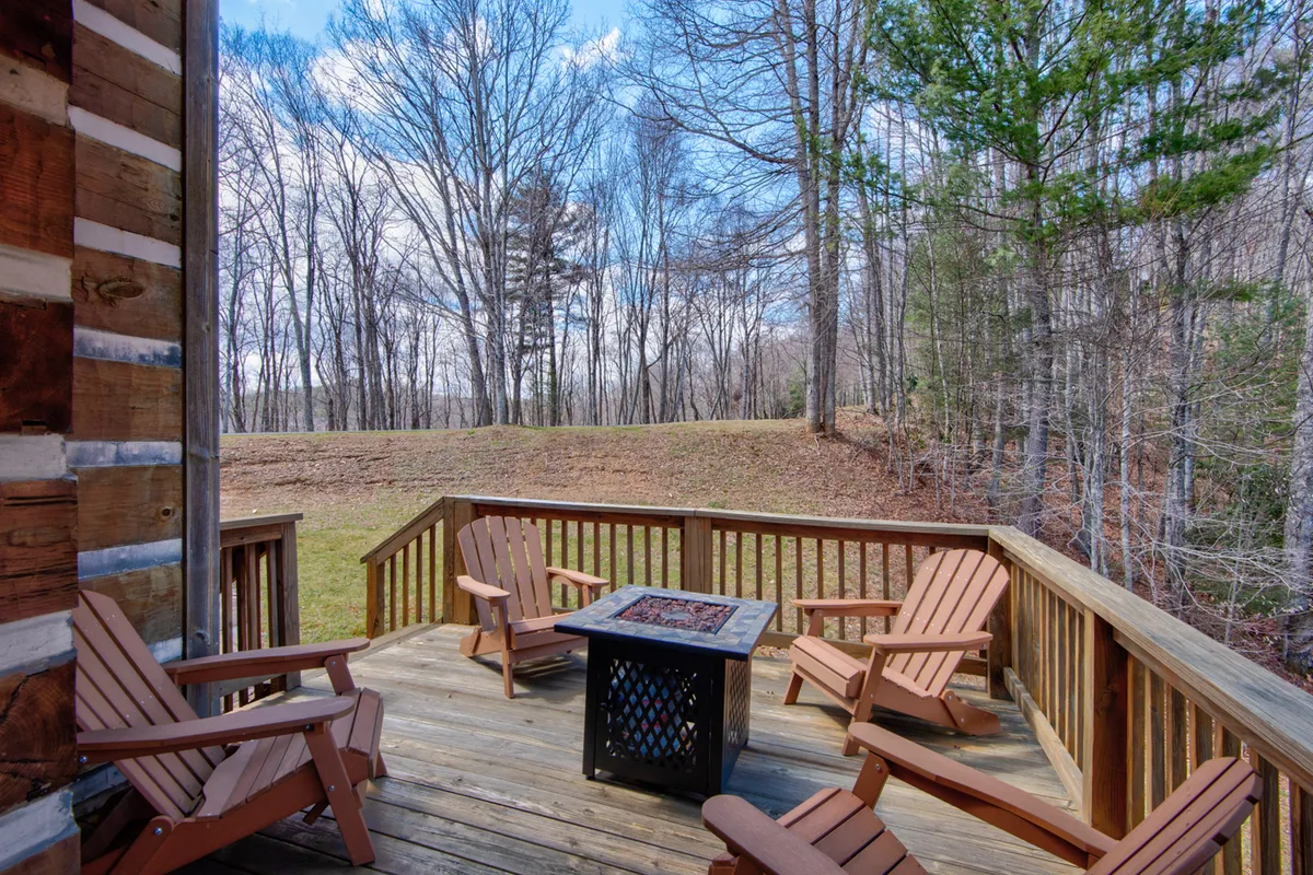 A spacious wooden deck set amidst tall trees, featuring several Adirondack chairs arranged around a gas fire pit. The deck faces a backdrop of lush greenery and distant mountains, creating an inviting space for outdoor gatherings and relaxation.