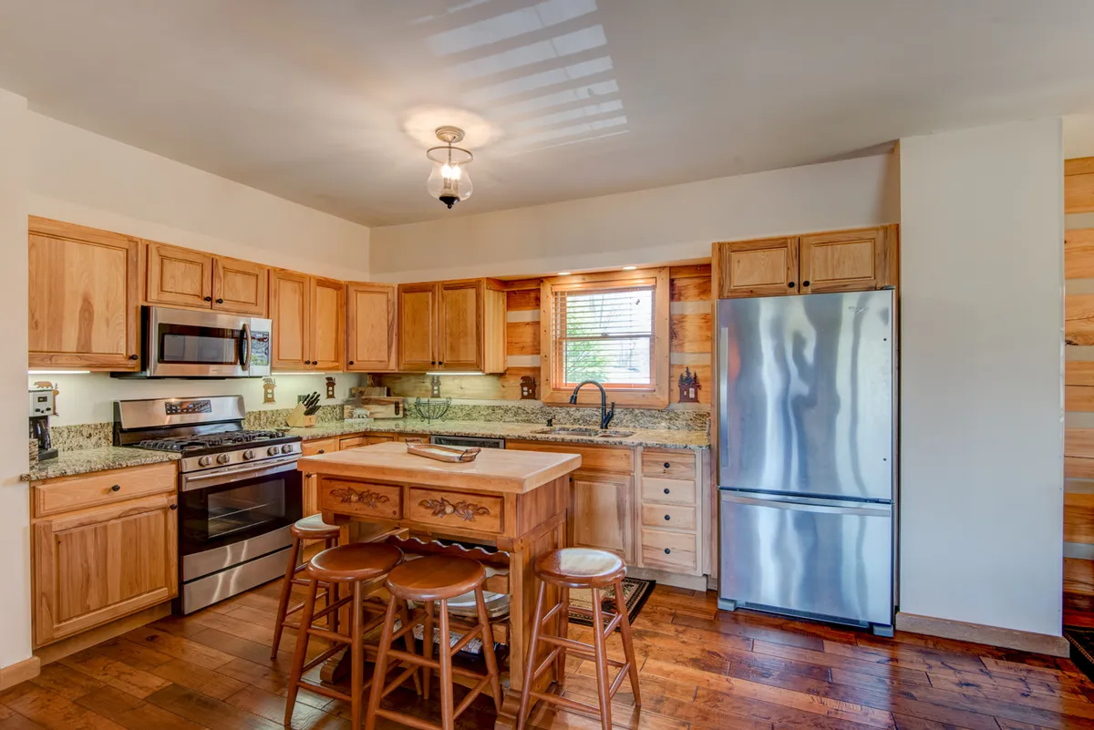 A well-equipped kitchen featuring wooden cabinetry, granite countertops, a central island with stools, and stainless steel appliances. Natural light streams through a window, highlighting an inviting space perfect for cooking and entertaining.