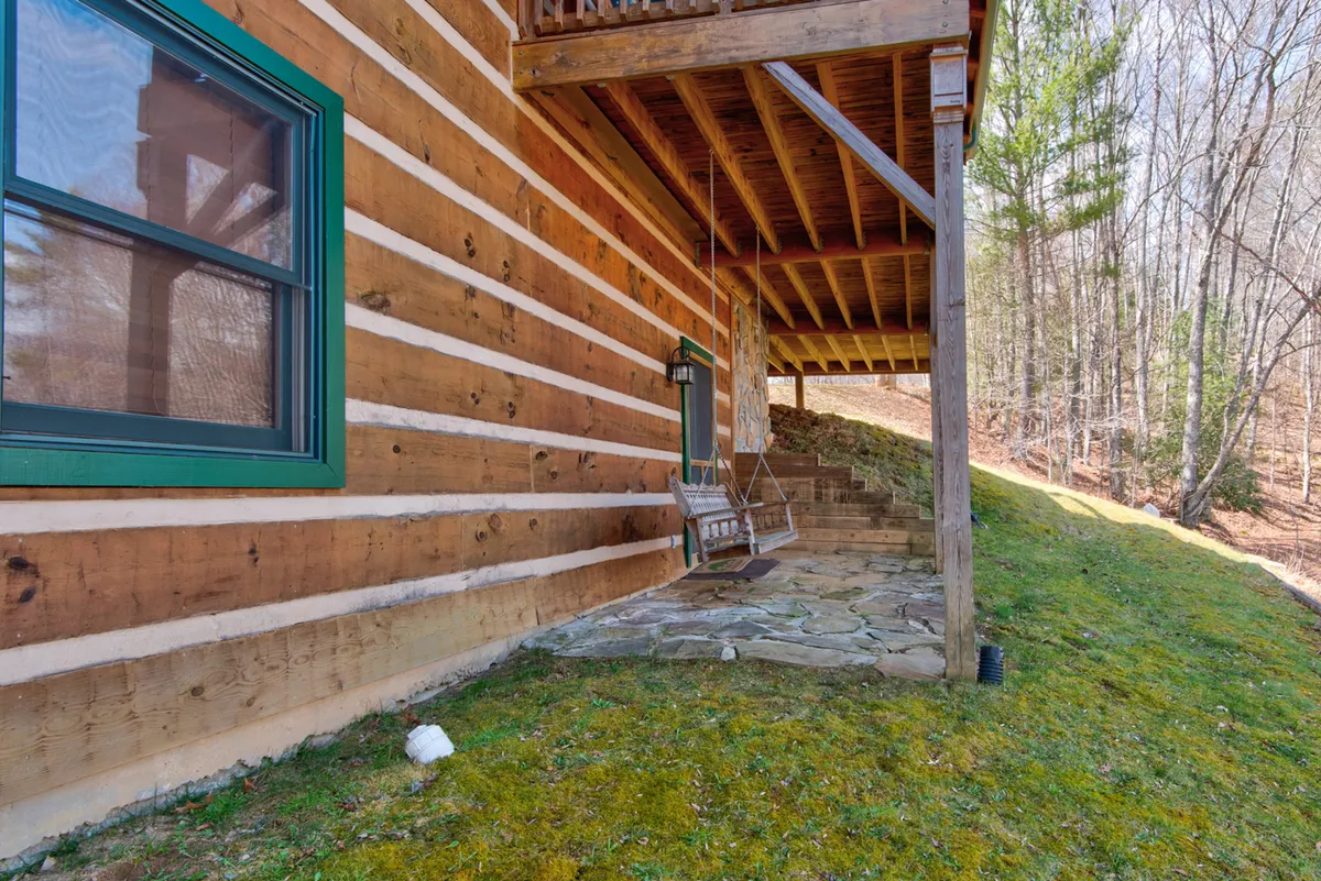 A charming view of the cabin's porch featuring a swing, stone pathway, and lush greenery. The porch swing invites moments of relaxation, whether watching the sunset, reading a book, or sharing a quiet conversation with family. The setting is tranquil and ideal for outdoor leisure, enhancing the theme of gathering and togetherness in nature.