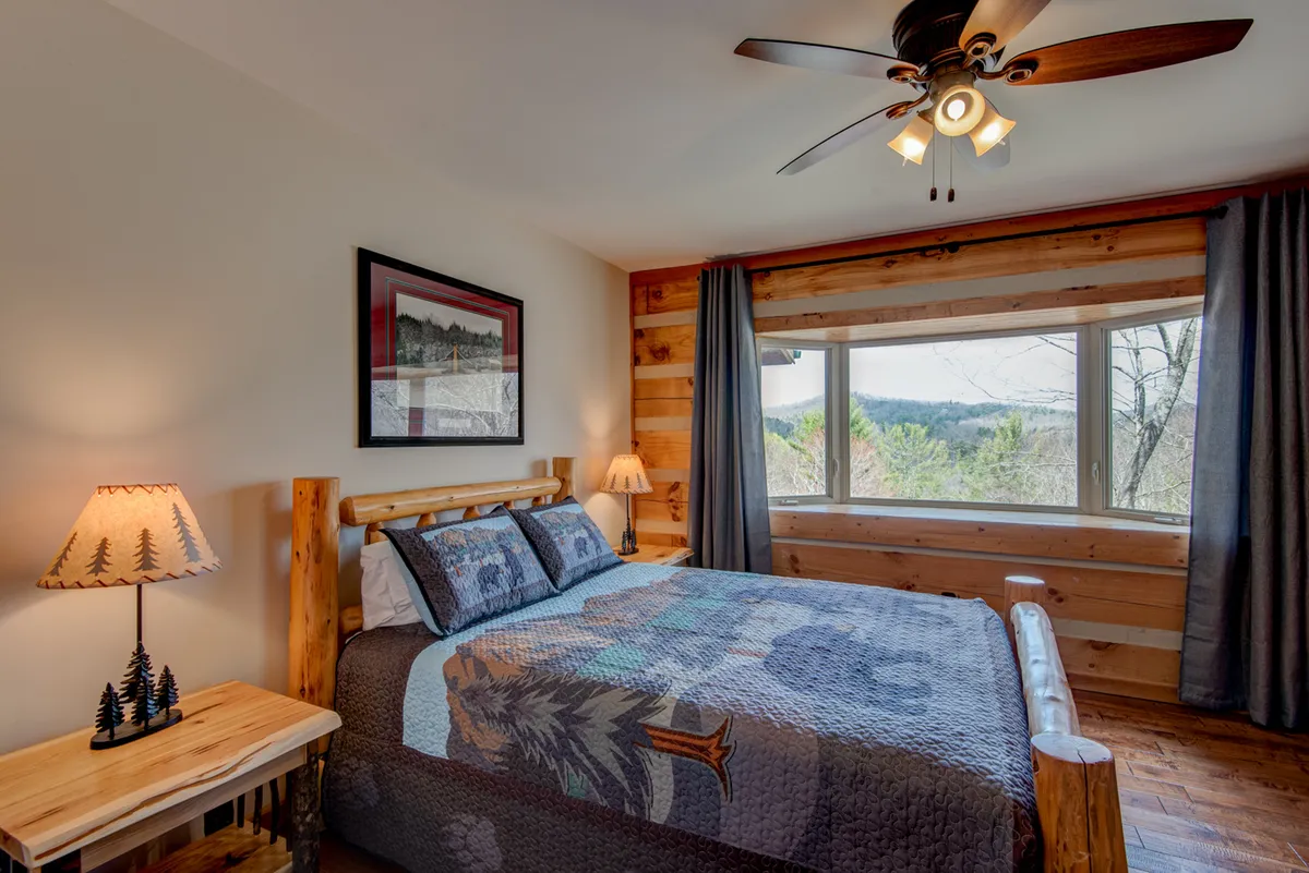 A sunlit bedroom features a bed with a patterned quilt, large window looking out to scenic views, wooden furniture, and a decorative lamp on the nightstand. The ambiance is serene and inviting, ideal for restful sleep or enjoying the tranquil landscape from bed.