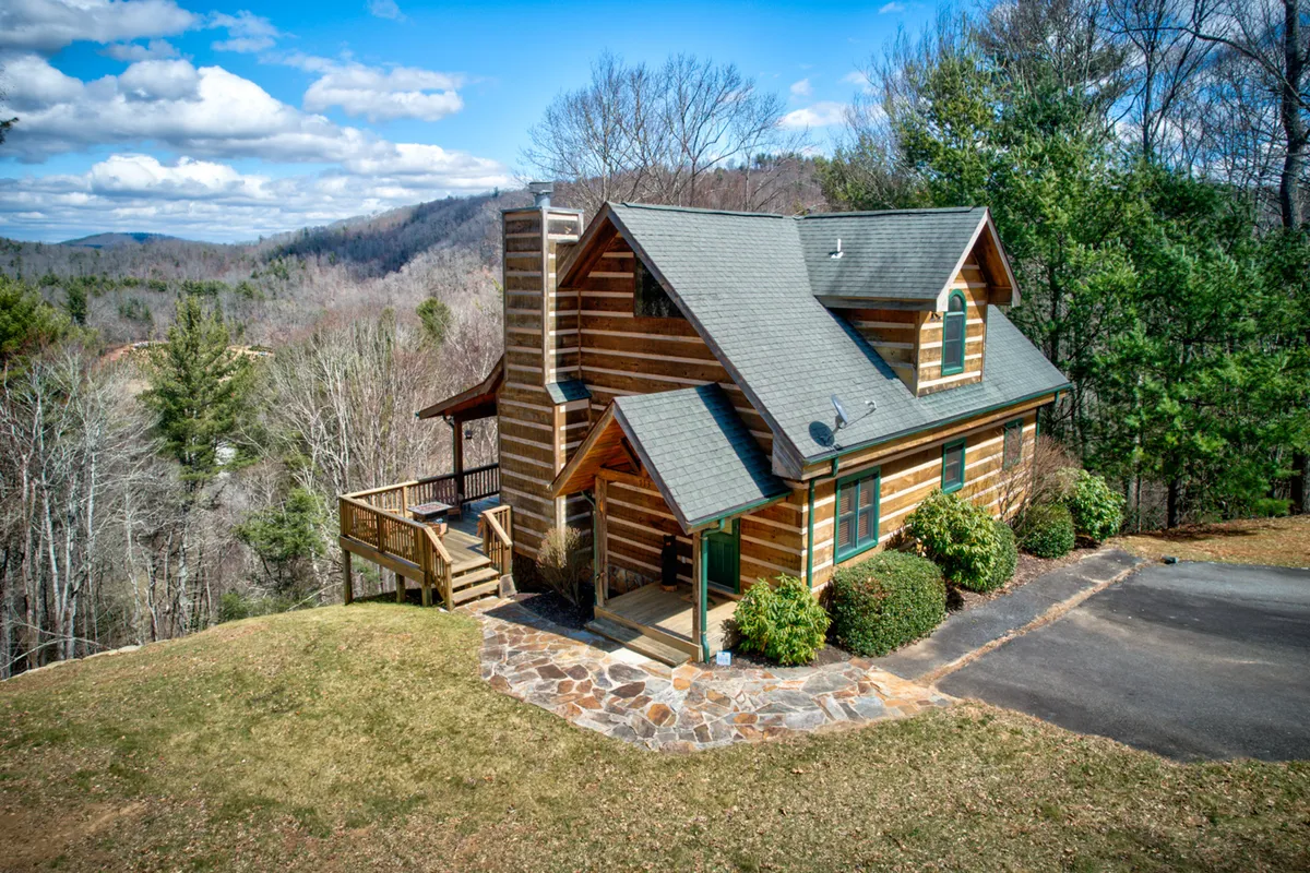 A classic log cabin is framed by a stone pathway and tall trees, suggesting inviting trails and easy movement between the cabin and the surrounding natural world. The dappled sunlight and peaceful atmosphere evoke mornings or evenings spent gathering outdoors before or after a river adventure.