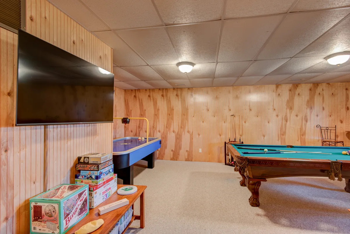 A bright, open game room equipped with both a pool table and an air hockey table, board games on the table, and a TV in clear view. The setting suggests fun, laughter, and multi-generational enjoyment, aligning with the room’s purpose as described.