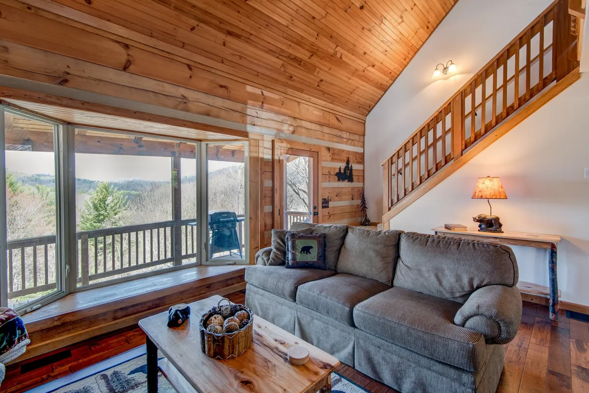 This interior shot features large windows offering sweeping mountain views, a comfortable couch, rustic wooden coffee table, and a wooden staircase. The warm, inviting atmosphere invites guests to relax with a book or enjoy scenic vistas from the comfort of the loft, aligning with the section's focus on cozy interiors and mountain panoramas.