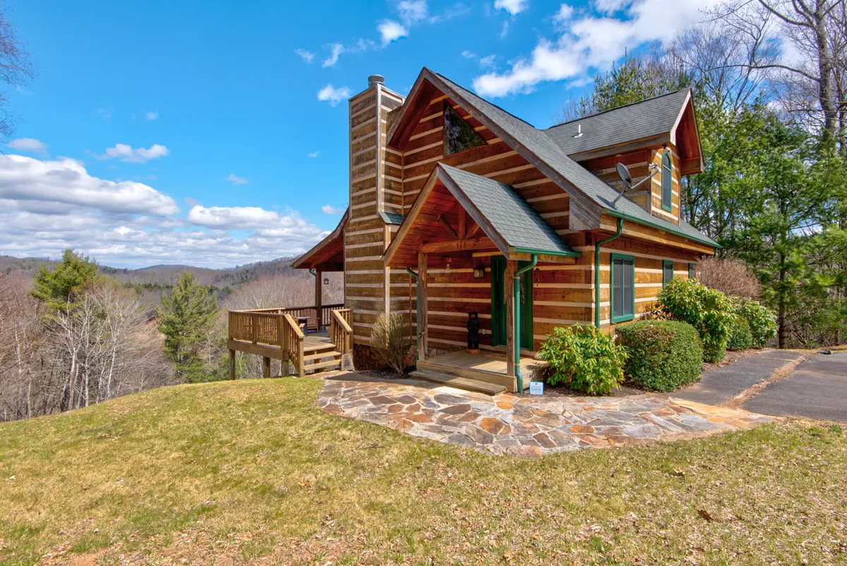 A charming three-story log cabin nestles in a peaceful mountain landscape, with a stone pathway leading up to a spacious deck perfect for soaking in sunset views. Surrounded by nature, the cabin promises a cozy, serene escape with ample outdoor space for relaxation and gathering.