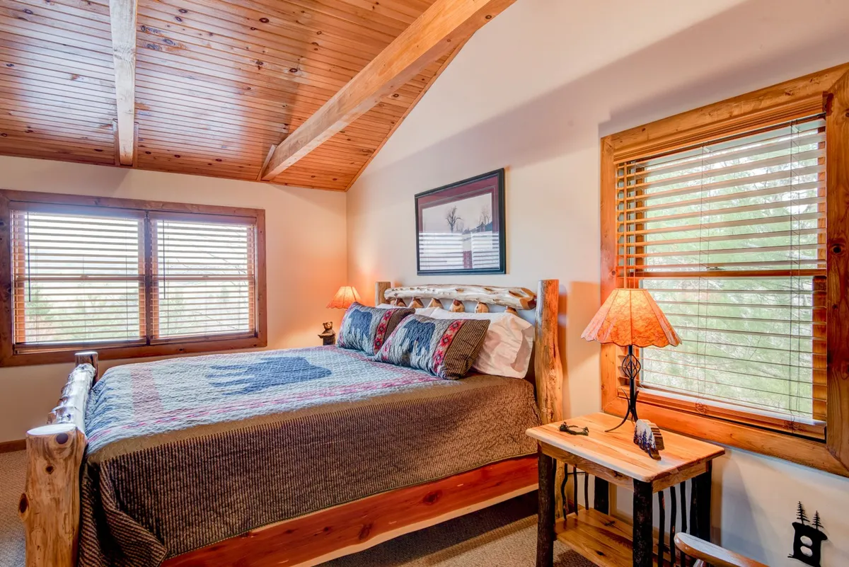A warmly decorated bedroom with a log bed, decorative pillows, wooden beams, soft lighting from bedside lamps, and windows letting in natural light. The space feels spacious, romantic, and connected to the outdoors.