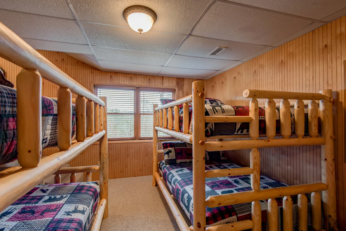 A cozy bunk room with wood-paneled walls, four queen-over-queen bunk beds dressed in plaid bedding, and windows that let in morning light. The room is playful and warm, perfect for group or family stays.