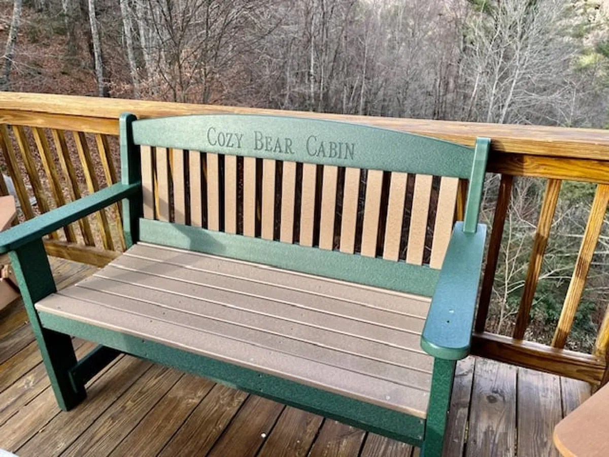 A tranquil bench labeled 'Cozy Bear Cabin' is nestled at the forest's edge, surrounded by lush trees and greenery. The forest floor is dotted with sunlight and seasonal flora, emphasizing a peaceful, natural setting perfect for unwinding after a day outdoors.