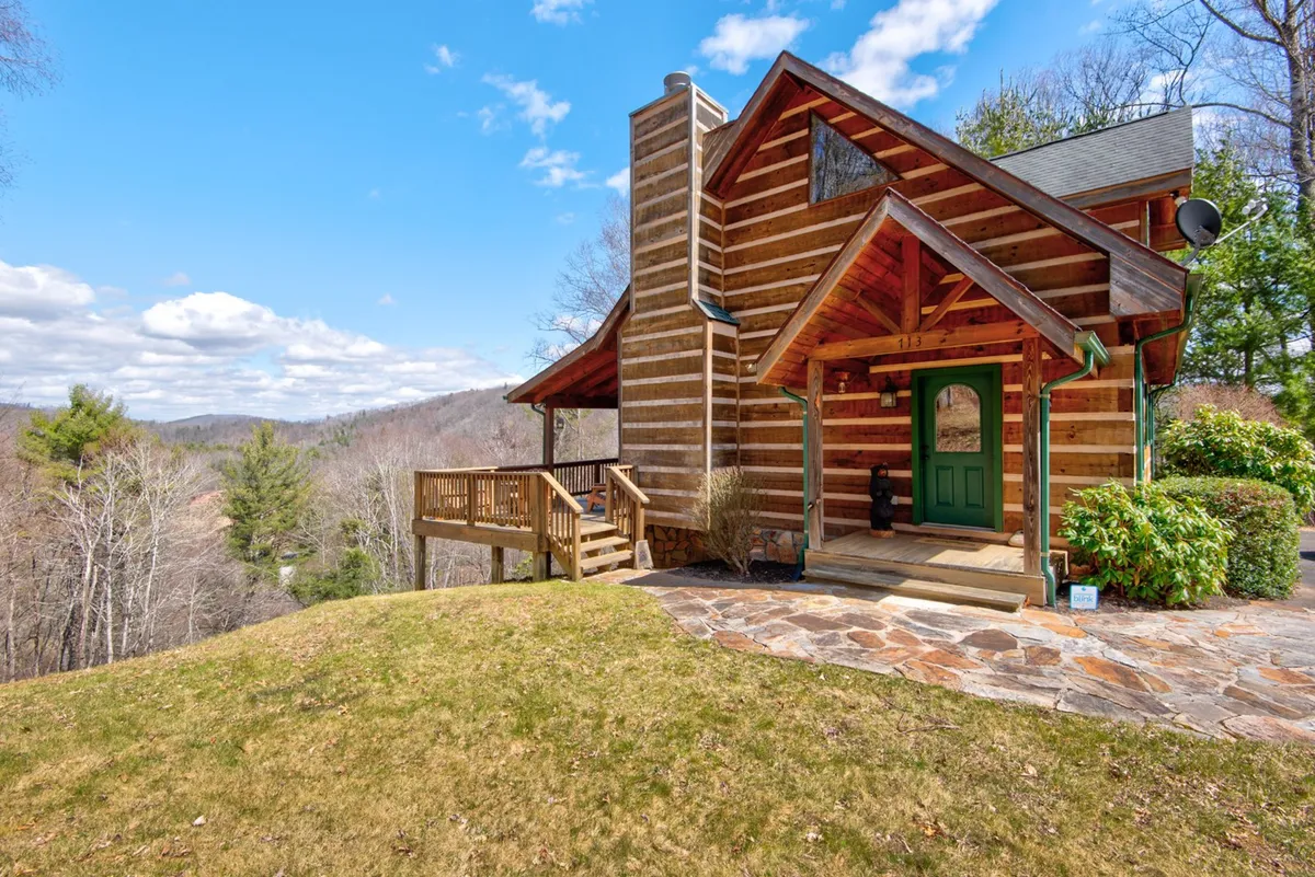 A welcoming log cabin exterior with a green front door and a stone pathway, surrounded by natural landscaping. The porch invites guests to relax and take in the mountain surroundings, suggesting easy access to trails and the outdoors.